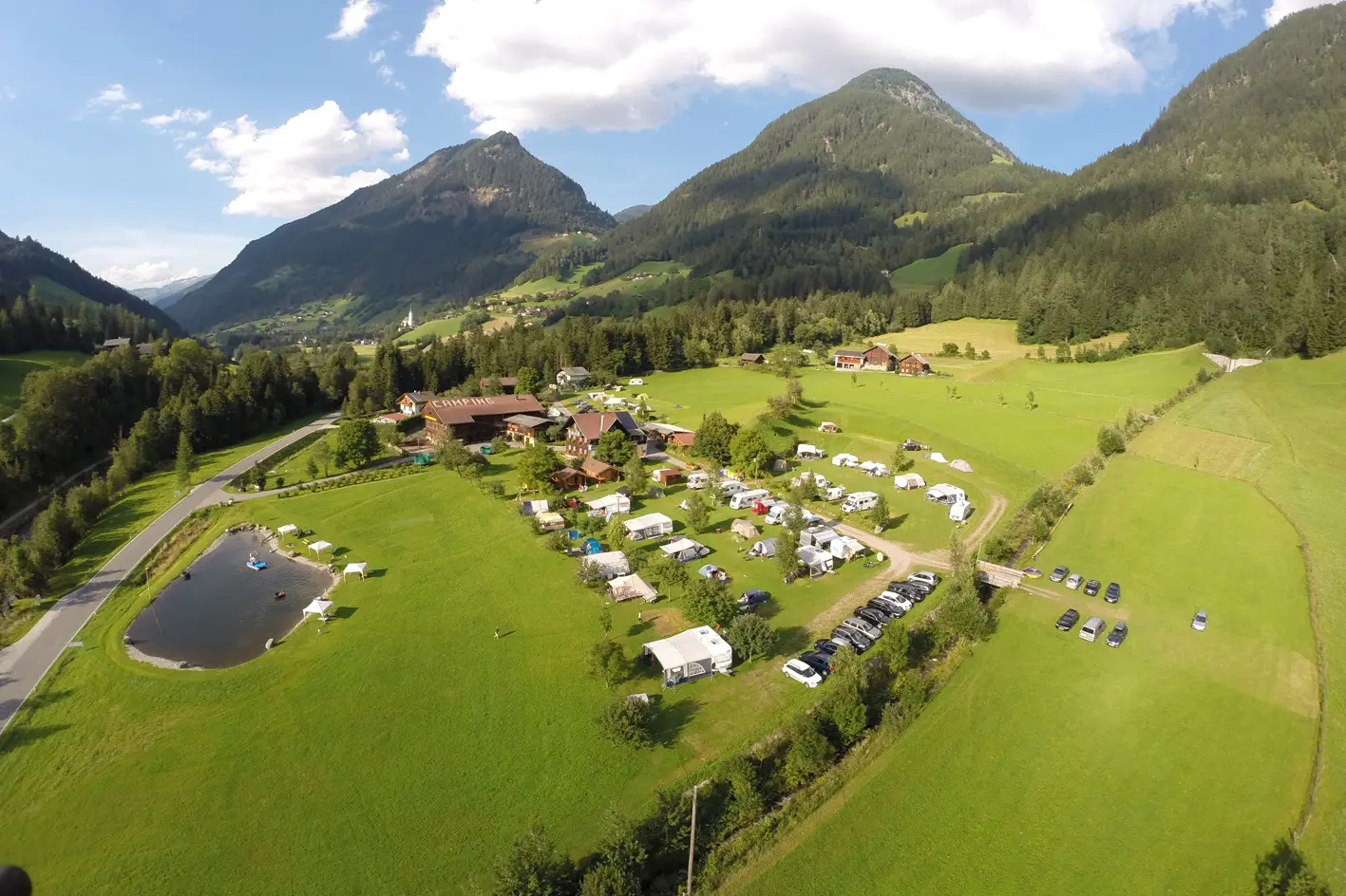 Camping Lindlerhof | Luftaufnahme eines üppig grünen Talcampingplatzes im Mölltal, Kärnten Urlaub, mit verstreuten Zelten und Wohnwagen. Auf der linken Seite ist ein kleiner Teich und in der Nähe sind Autos geparkt. Umgeben von Bergen und Wäldern unter einem blauen Himmel mit flauschigen Wolken.