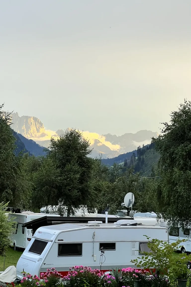 Camping Lindlerhof | Ein malerischer Campingplatz mit einem weißen Wohnwagen, umgeben von Grün und blühenden Büschen im Herzen des Mölltals in Kärnten. Im Hintergrund sind schneebedeckte Berge unter einem klaren Himmel zu sehen, die eine ruhige und malerische Landschaft bilden, die perfekt für einen unvergesslichen Urlaub ist.