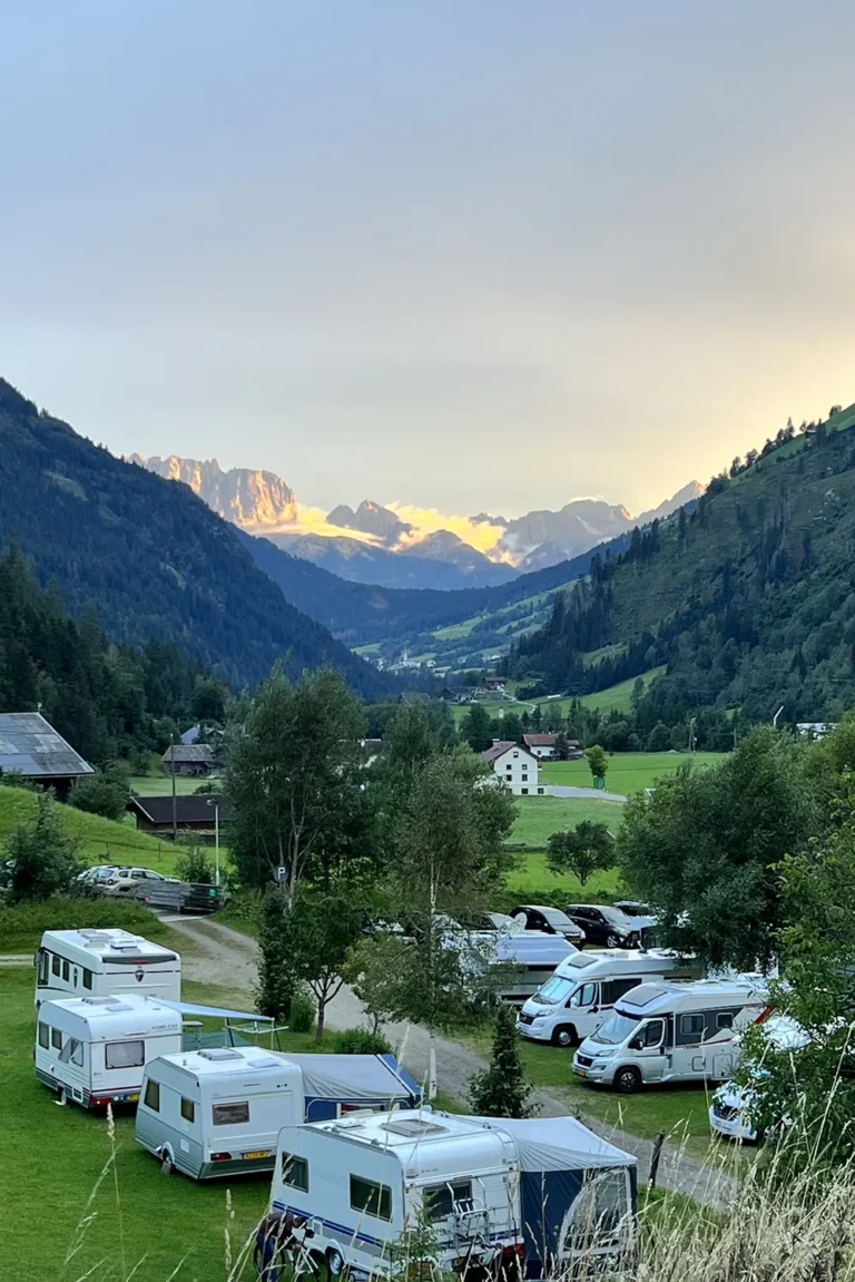 Camping Lindlerhof | Ein malerisches Tal im Mölltal in Kärnten, mit Wohnmobilen auf grünem Gras, umgeben von üppigen Bäumen und Bergen. Schneebedeckte Gipfel erheben sich majestätisch im Hintergrund unter einem teilweise bewölkten Himmel bei Sonnenuntergang – ein perfekter Ort für einen unvergesslichen Urlaub.