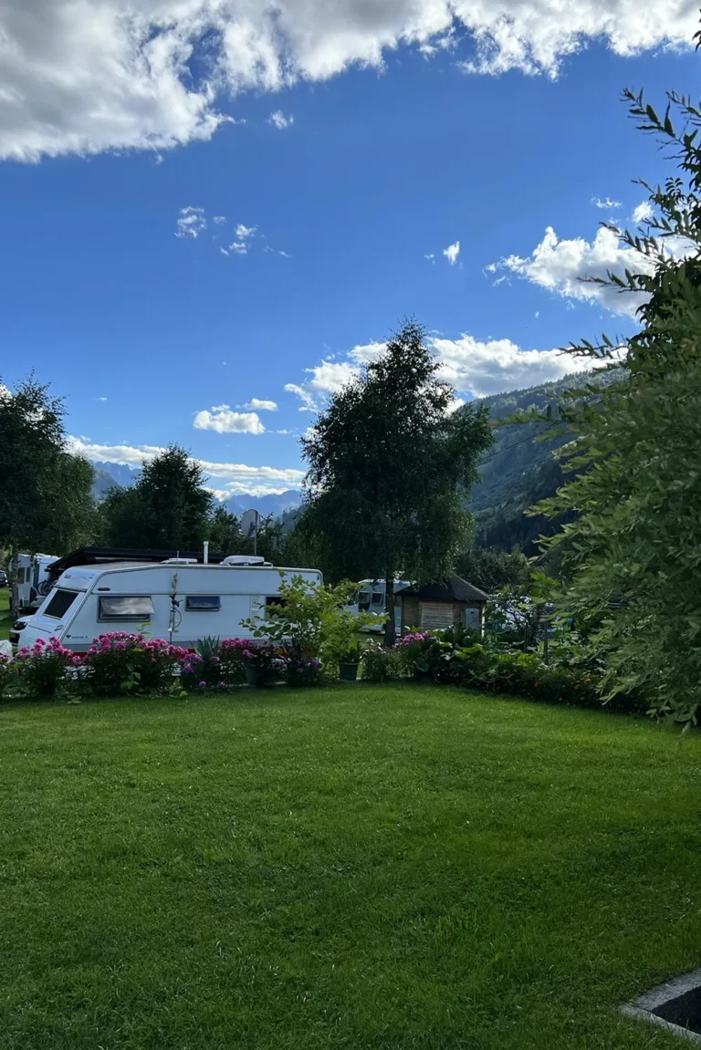 Camping Lindlerhof | Ein weißer Camper steht auf einer üppigen grünen Wiese im Mölltal in Kärnten, umgeben von Bäumen und leuchtenden Blumen. Die majestätischen Berge erheben sich stolz im Hintergrund unter einem blauen Himmel mit vereinzelten Wolken und machen es zu einem idyllischen Ort für einen unvergesslichen Urlaub.