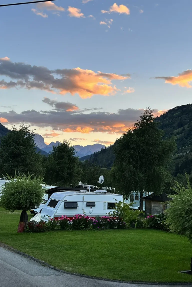 Camping Lindlerhof | Ein Wohnwagen steht auf einer üppigen grünen Wiese im Mölltal in Kärnten, umgeben von Bäumen und leuchtenden Blumen. Im Hintergrund sorgen Berge und ein Sonnenuntergangshimmel mit orangefarbenen Wolken für eine malerische Urlaubsszene.