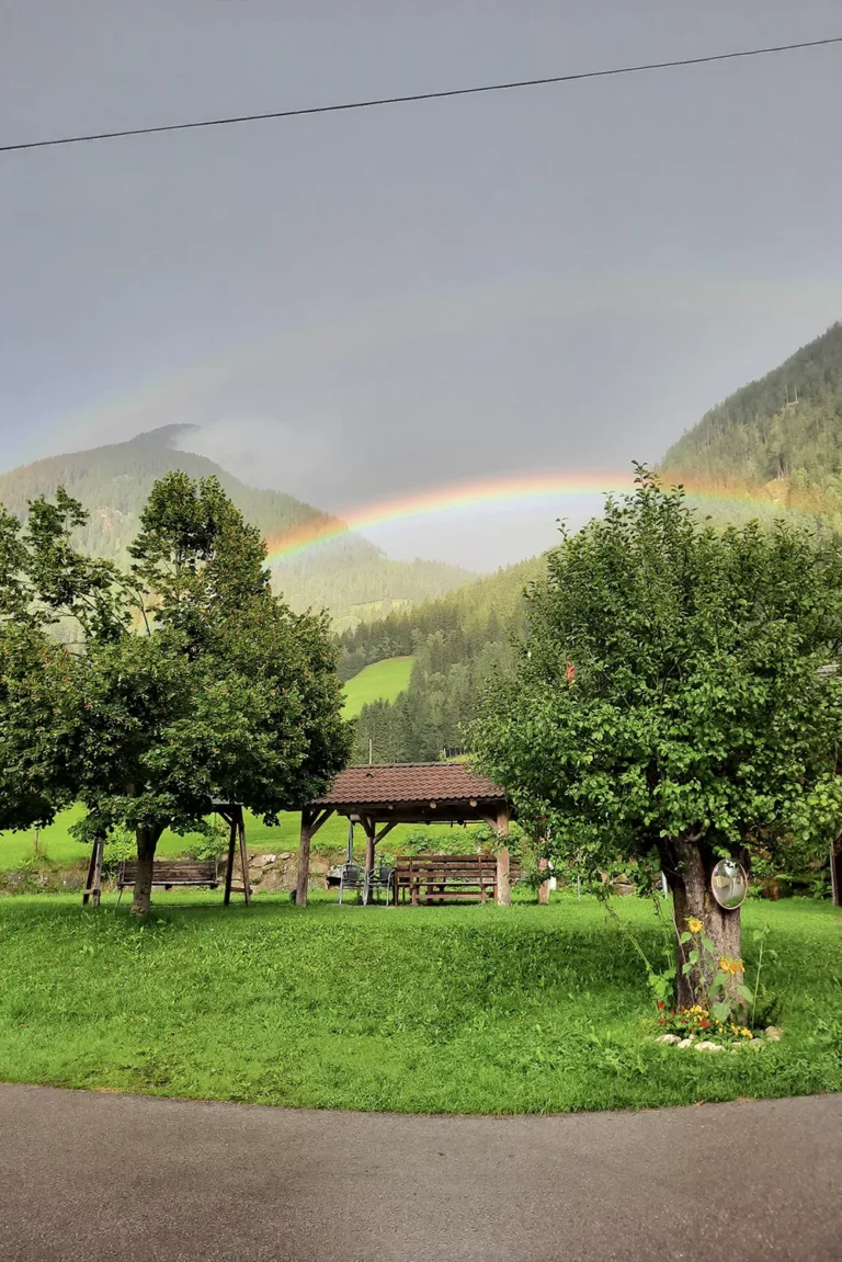 Camping Lindlerhof | Ein leuchtender Regenbogen wölbt sich über dem Mölltal in Kärnten mit üppigen grünen Hügeln und Bäumen. Darunter steht ein Holzunterstand auf einer Wiese und fängt die ruhige Atmosphäre dieses perfekten Urlaubsortes ein. Der teilweise bewölkte Himmel verleiht der malerischen Landschaft eine friedliche Atmosphäre.