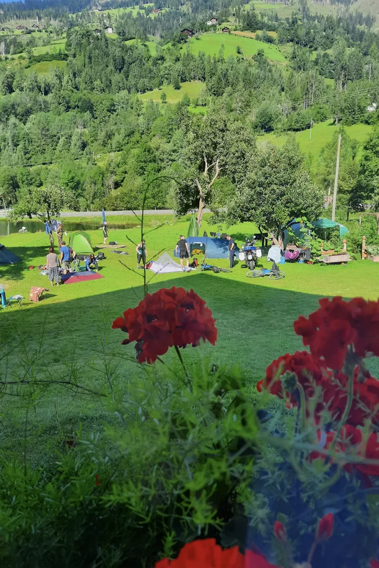 Camping Lindlerhof | Ein malerischer Campingplatz auf einem üppig grünen Hügel im Mölltal Kärnten, mit mehreren Zelten und herumlungernden Menschen. Im Vordergrund sind leuchtend rote Blumen zu sehen. Im Hintergrund sind bewaldete Hügel unter einem klaren blauen Himmel zu sehen, perfekt für einen erholsamen Urlaub.