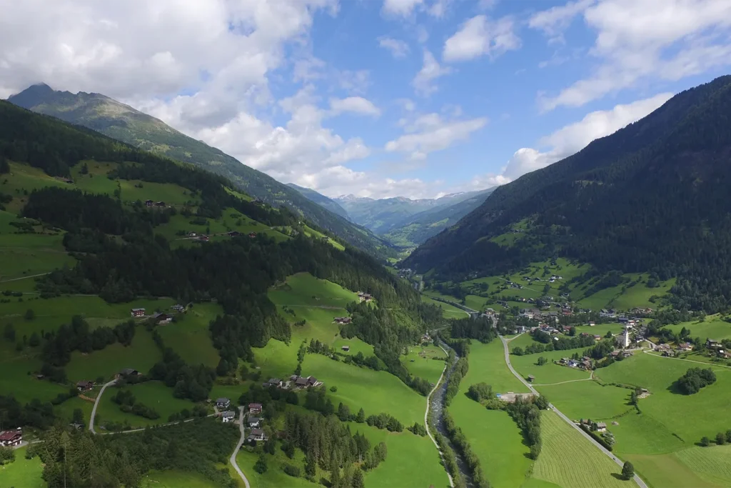 Camping Lindlerhof | Eine malerische Luftaufnahme des üppig grünen Mölltals in Kärnten mit verstreuten Häusern. Ein Fluss schlängelt sich durch die Landschaft, flankiert von bewaldeten Hügeln und Bergen. Der Himmel ist teilweise bewölkt, was dieser ruhigen ländlichen Szene Tiefe verleiht, perfekt für einen unvergesslichen Urlaub.