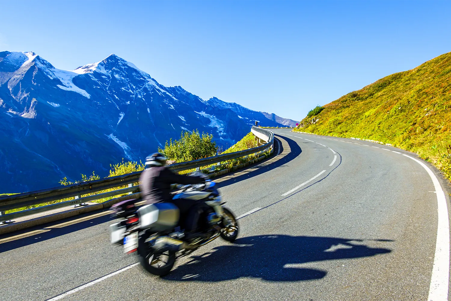 Camping Lindlerhof | Ein Motorradfahrer fährt eine kurvenreiche Bergstraße im Mölltal in Kärnten entlang, mit leuchtend grünen Hügeln auf der einen Seite und hoch aufragenden schneebedeckten Gipfeln auf der anderen. Der klare blaue Himmel verstärkt diese atemberaubende Aussicht, perfekt für einen unvergesslichen Urlaub.