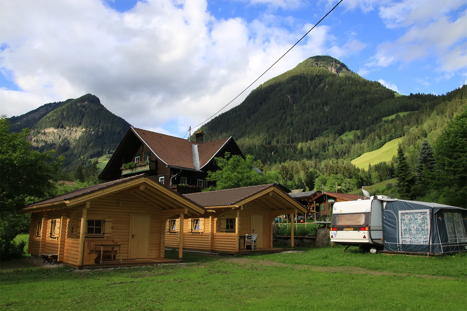 Camping Lindlerhof | Eine malerische Aussicht auf Chalets und einen Wohnwagen auf einer Wiese im Mölltal, Kärnten, umgeben von üppigen grünen Bäumen und hohen Bergen unter einem teilweise bewölkten Himmel. Darüber verläuft eine Stromleitung, die dem rustikalen Urlaubsambiente einen Hauch von Modernität verleiht.